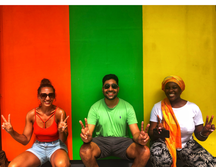 Three students holding up peace signs sitting in front of a wall painted in red, green, and yellow.