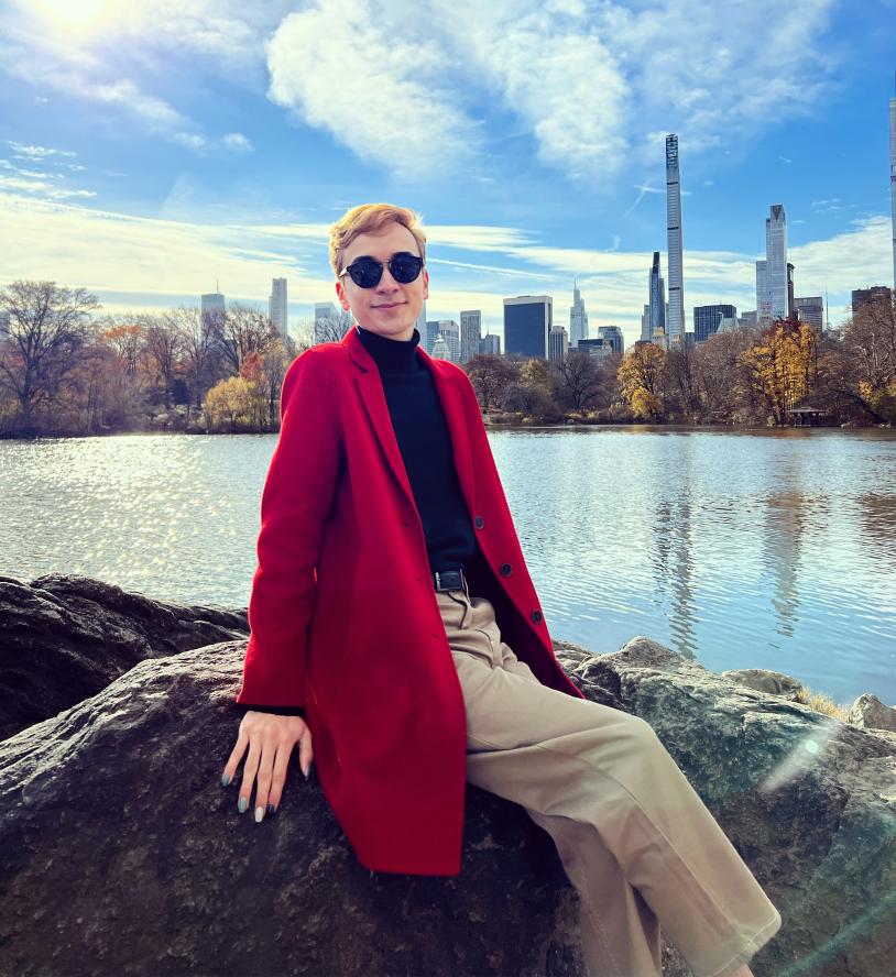 joel smiles at the camera sitting on a boulder in front of a small body of water and city skyline in Central Park, New York City. She is wearing tan pants, a black turtle neck, a long red coat, and black sunglasses - and their multi colored nails are on display.