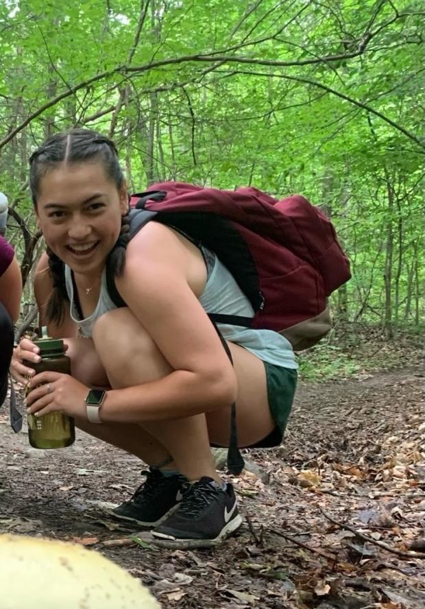 Coordinator Lina Fang smiling and squatting in the woods with a backpack on 