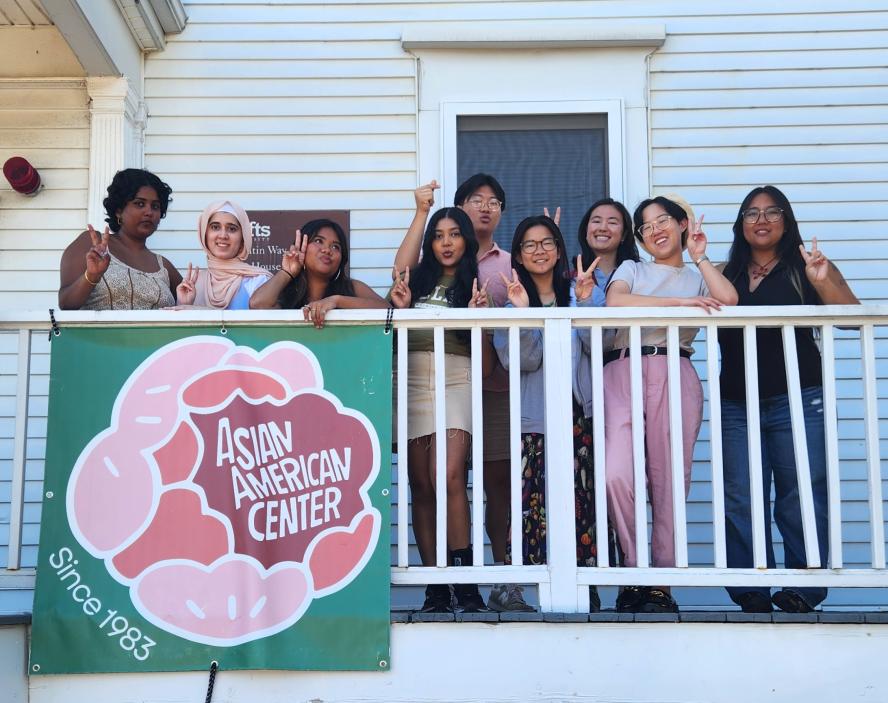 Nine individuals pose with peace signs in on a white porch behind the Asian American Center logo banner.